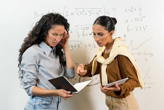 Cute Oriental Girls Solving a Problem. Students are discussing, looking into a notebook and not understanding math equation. Schoolgirls of the Higher School stand near the blackboard