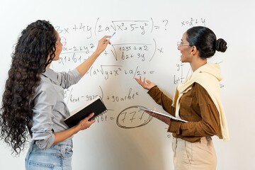 Two Middle Eastern female students stand near the blackboard and show equations. Woman jointly solves tasks holding a tablet and a tablet in her hands. Girls with curly hair do education