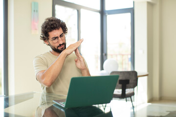 young adult bearded man with a laptop looking serious, stern, angry and displeased, making time out sign