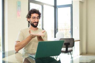 young adult bearded man with a laptop smiling happily with friendly, confident, positive look, offering and showing an object or concept