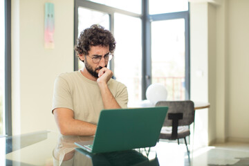 young adult bearded man with a laptop feeling serious, thoughtful and concerned, staring sideways with hand pressed against chin