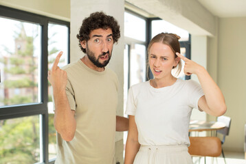 young adult couple feeling confused and puzzled, showing you are insane, crazy or out of your mind