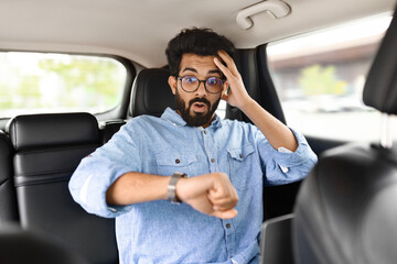 Stressed eastern man looking at watch, touching head in car