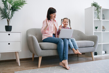 Obraz premium Brunette female in jeans and smiling child with braids waving hello while looking at laptop screen on knees at home. Cheerful ladies greeting relatives during online video call using internet app.
