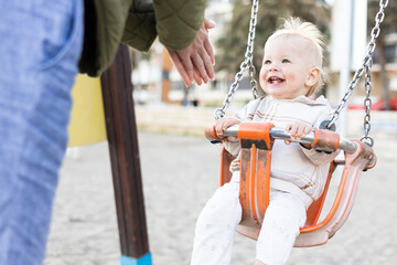 Mother pushing her cheerful infant baby boy child on a swing on sandy beach playground outdoors on nice sunny cold winter day in Malaga, Spain