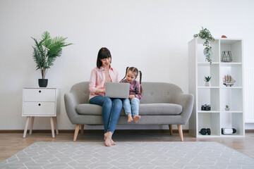 Obraz premium Pretty caucasian mother and daughter in cozy outfits holding portable computer while resting on couch at home. Happy family having cinema evening with ordering pizza online at sunny weekend.