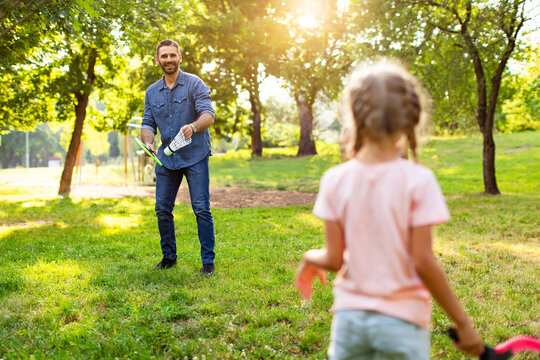 Rear view of middle aged man holding badminton racket and big shuttlecock while playing with his daughter in the park
