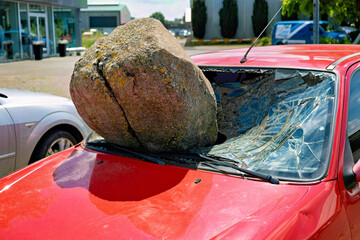 A large round stone rests on the hood of a red car with a shattered windshield
