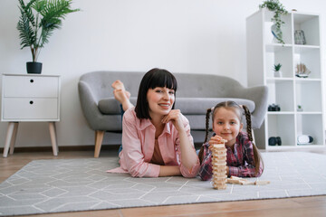 Portrait of beautiful young mother and little daughter relaxing on warm floor behind wooden toy tower indoors. Happy family enhancing relationship bonds by having game night at home together.