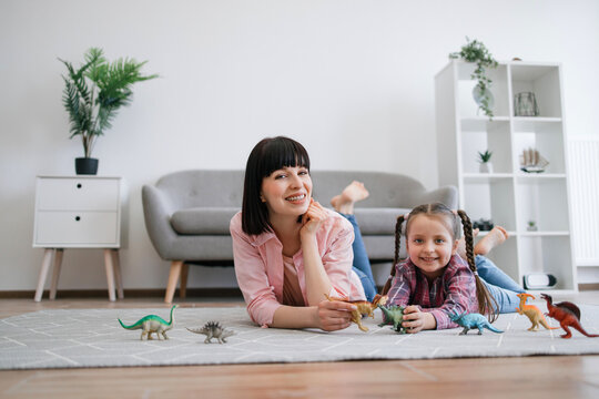 Portrait of mother-daughter duo posing with amazing toys of extinct reptiles in hands while lying comfortably on floor. Smiling caucasian ladies gaining knowledge about different animals and habitats.