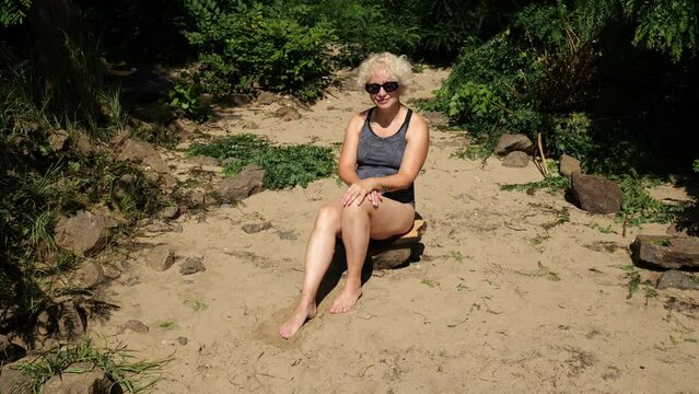 A beautiful fifty year old woman in a dark swimsuit gets up from the sand in the jungle