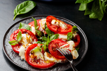 Caprese Salad. Tomato and mozzarella slices with basil leaves with fork on dark plate. Traditional Italian food