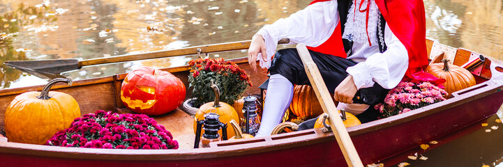 banner young man dressed as gondolier in gondola boat decorated with pumpkins on pond in autumn park celebrates Halloween and having fun, concept of Halloween carnival or costume party - obrazy, fototapety, plakaty