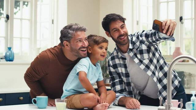 Same sex family with two Dads in kitchen at home with son sitting on counter posing for selfie on mobile phone - shot in slow motion