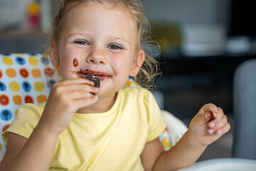 Little girl with blond hair eating homemade chocolate with dirty mouth and hands in home kitchen