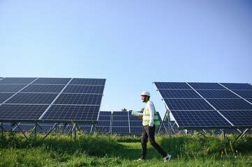 Portrait of Young indian male engineer standing near solar panels, with clear blue sky background,...