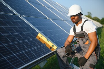 African american man in white helmet and grey overalls standing among rows of solar panels