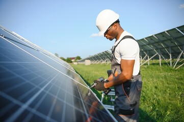 African american man in safety helmet and glasses tighten nuts on solar panels with screwdriver....
