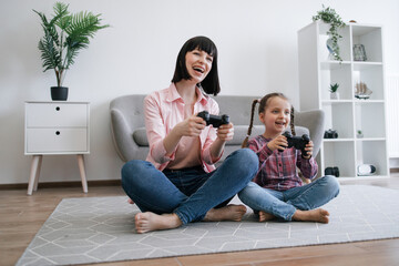 Obraz premium Delighted female parent and daughter holding black joypads while relaxing on room floor with legs crossed. Overjoyed family members playing video games for competition and computer learning.