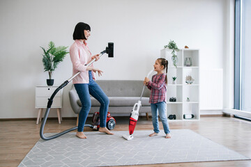 Excited mother using vacuum cleaner as imaginary guitar while daughter pretending rock singer with...