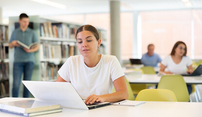 Obraz premium Positive young lady in casual clothes working remotely using a computer in a quiet library