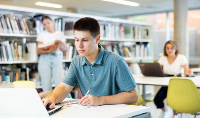 Portrait of positive young man with laptop and book in public library