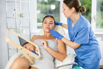Woman cosmetologist examines patients nasolabial area after lifting procedure and gently palpates touches lips mouth zone with finger