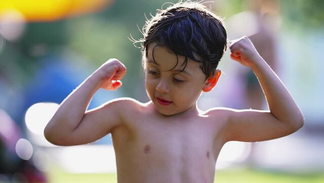 One adorable little boy flexing muscles sitting on grass outside after swimming at the pool during warm summer day. Child shows tiny biceps