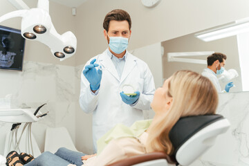 Portrait male dentist in protective mask showing aligners to female patient sitting in dental chair