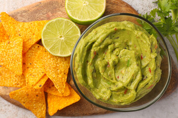 Wooden board with bowl of delicious guacamole, nachos chips and lime on white tiled table, top view