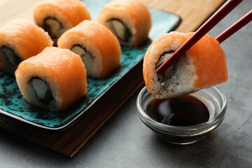 Dipping tasty sushi roll with salmon into soy sauce on grey table, closeup