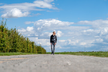 Jeune homme en balade sur une petite route de campagne sous un ciel bleu avec quelques nuages - obrazy, fototapety, plakaty