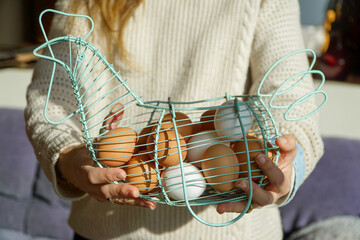 Up-close view of a woman's hands gently cradling a whimsical blue chicken-shaped basket filled with a delightful assortment of fresh, organic eggs.