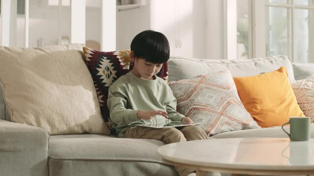 five-year-old little asian boy sitting on couch at home playing computer game with digital tablet