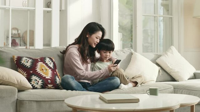 young asian mother and five years old son sitting on couch at home looking at mobile phone together