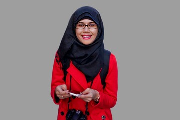 Close up portrait of smiling young Muslim woman wearing black hijab and red jacket with DSLR camera hanging from neck holding phone. Happy and cheerful expression.
