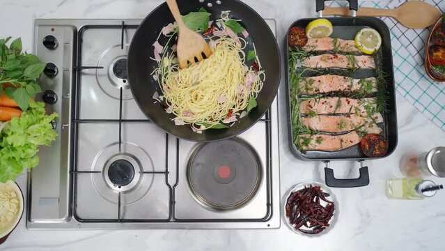 Asian woman cooking healthy food fried pasta and salmon steak on cooking pan in the kitchen at home. Attractive girl preparing food for dinner party celebration meeting with friend on holiday vacation