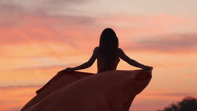 A young woman with long hair in a red dress developing in the wind epic walks barefoot through the sand dunes of the desert against the backdrop of the setting sun