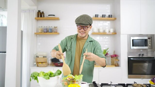 4K Portrait of Asian man cooking healthy food salad on modern kitchen island stove at home. Happy guy preparing food meal for dinner party celebration meeting with friends on holiday vacation.