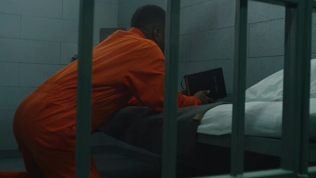 African American prisoner in orange uniform kneels near the bed, prays to God in prison cell with Bible. Male criminal serves imprisonment term in jail or detention center. Concept of faith in God.