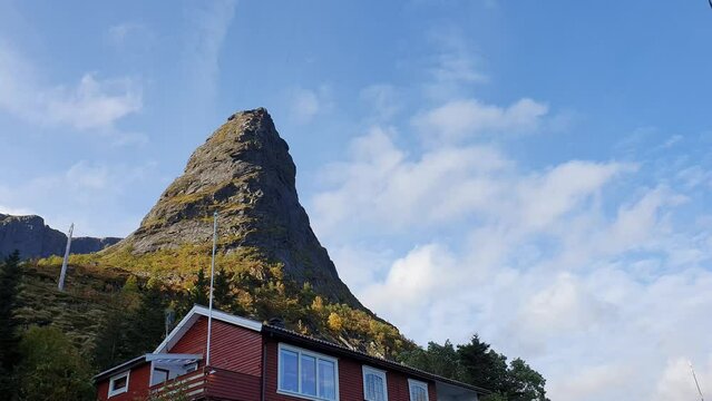 View over a red norwegian hut at the foot of a tippy mountain against a blue sky.