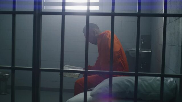 African American prisoner in orange uniform sits on the bed, reads Bible in prison cell. Male criminal serves imprisonment term for crime in jail or detention center. Faith in God. View through bars.