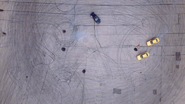 An Overhead Aerial View of a Black Drift Car Burning Rubber on in an Asphalt Parking Lot Joined by Two Yellow Race Cars in High Horsepower Tandem Drifting