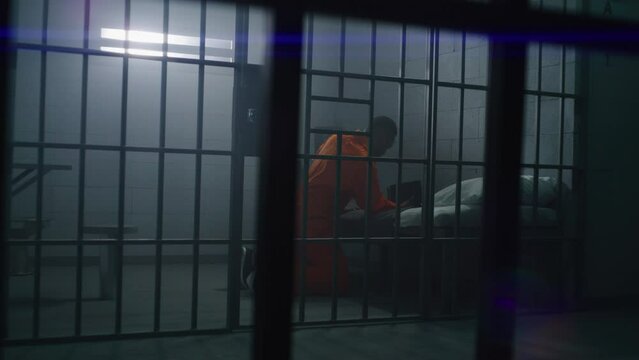 African American prisoner in orange uniform kneels near bed, prays to God in prison cell with Bible. Male criminal serves imprisonment term in jail or correctional facility. View through metal bars.