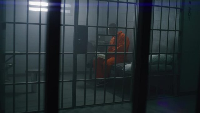 African American prisoner in orange uniform sits on the bed, reads Bible in prison cell. Prison officer walks, watches criminal. Inmate serves imprisonment term for crime in jail or detention center.