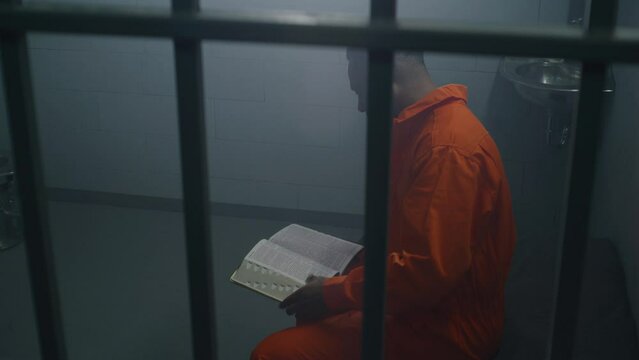 African American prisoner in orange uniform sits on bed behind bars, reads Bible in prison cell. Male criminal serves imprisonment term for crime in jail. Detention center or correctional facility.