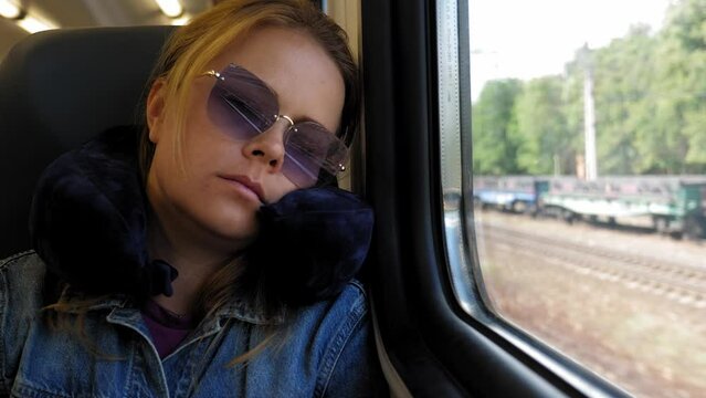 A tired young woman in sunglasses with a travel neck pillow sleeps on the train leaning on the window during a summer trip.