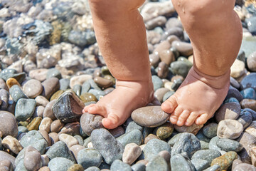 Infant boy standing on pebbles with wet feet on sunny beach. Child casting shadow explores pebbles on sunlit sea beach at seaside resort