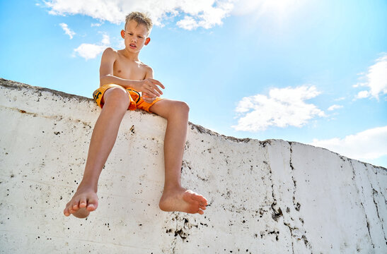 Preteen boy sits on sunny pier watching seaside against blue sky. Blond schoolboy spending summer vacation at sunny seaside resort