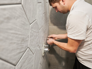 Male plumber doing plumbing works while working on bathroom renovation at home. Close up of man standing by the wall with ceramic tile and installing shower unit components in apartment.